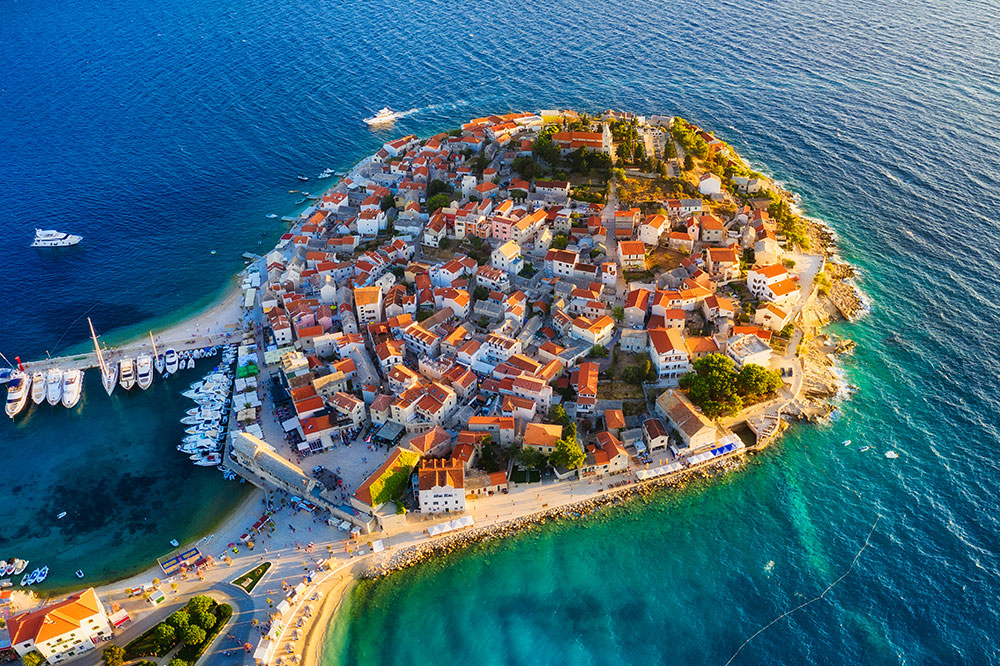 Widok z lotu ptaka na nadmorskie miasto z charakterystycznymi czerwonymi dachami otoczone przez turkusowe morze.