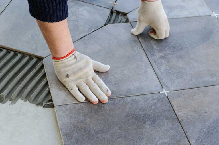 Osoba układająca szare płytki ceramiczne na podłodze, nosząca rękawice robocze.