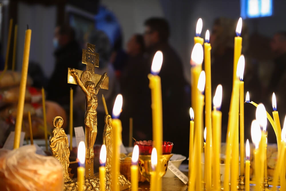 Świeczki zapalone w cerkwi, złoty krucyfiks na pierwszym planie, rozmyte postacie w tle.