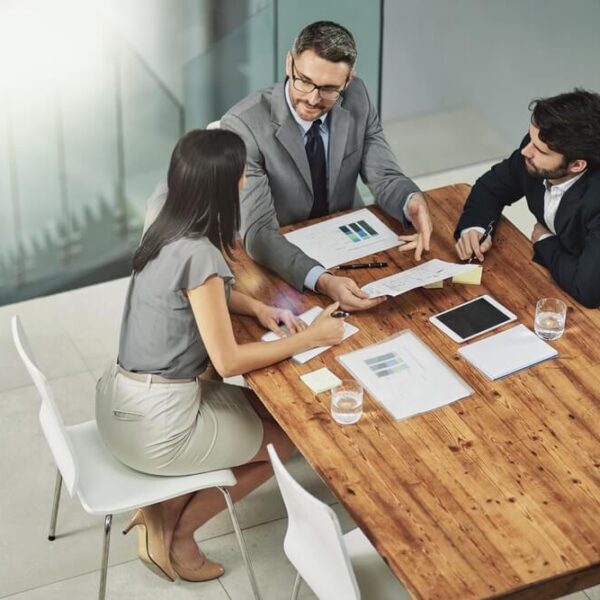 Trzy osoby siedzące przy drewnianym stole w biurze, dyskutujące nad dokumentami i urządzeniami elektronicznymi.