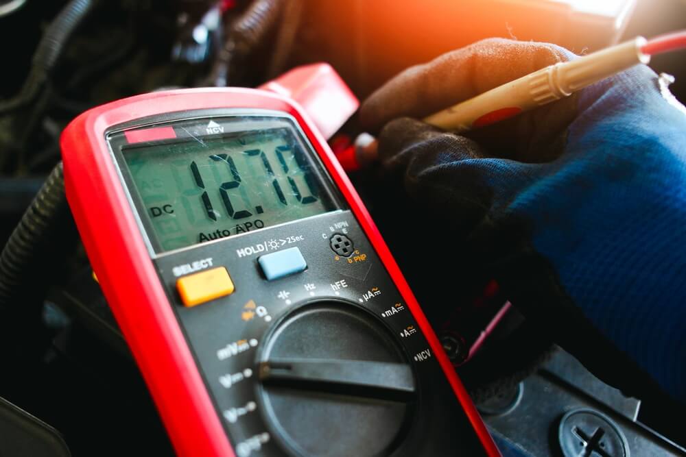 Multimetr z wyświetlaczem pokazującym wartość 12.7V, trzymany przez osobę w rękawicy.