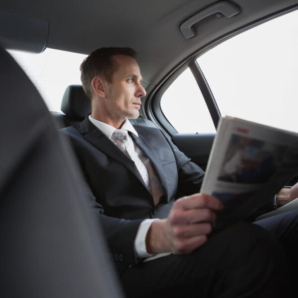 Mężczyzna w garniturze siedzi na tylnym siedzeniu samochodu, trzymając gazetę.
