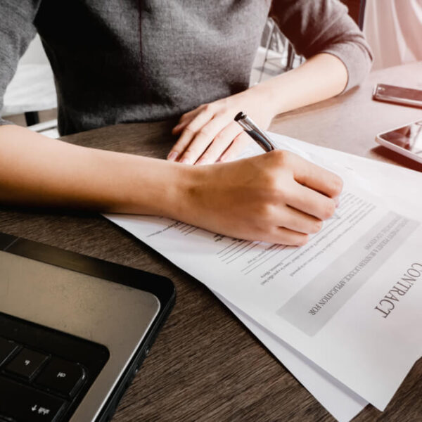 Osoba podpisująca dokumenty na stole obok laptopa, telefonu i tabletu.