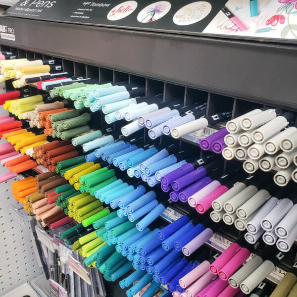 Stojak z kolorowymi pisakami Tombow ABT PRO, ułożonymi w szeregi według odcieni, od ciepłych barw po chłodne, na tle perforowanej ściany w sklepie artystycznym.