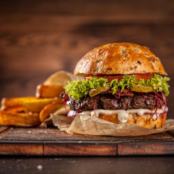 Burger z bułką z sezamem, wołowiną, sałatą, pomidorem i cebulą na drewnianej desce, obok frytki.