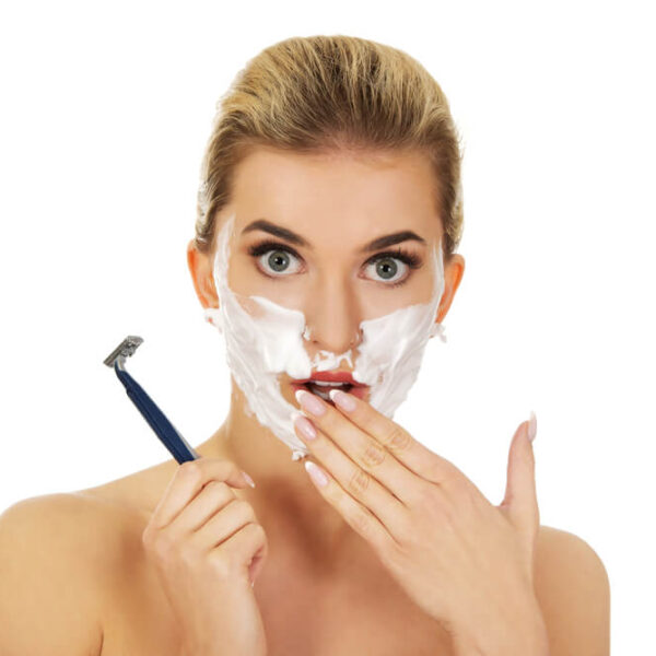 Kobieta trzymająca golarkę, z kremem do golenia na twarzy, wyglądająca na zaskoczoną.