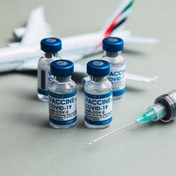 Kilka fiolek szczepionki na COVID-19 i strzykawka ustawione przed modelem samolotu na szarym tle.