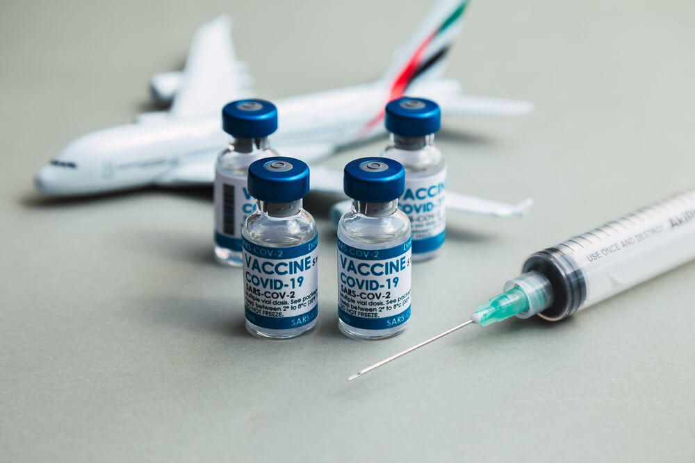 Kilka fiolek szczepionki na COVID-19 i strzykawka ustawione przed modelem samolotu na szarym tle.