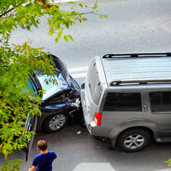Widok z góry na stłuczkę drogową, gdzie czarny samochód osobowy zderzył się z tyłu szarego SUV-a na ulicy otoczonej drzewami. Obok pojazdów stoi mężczyzna w niebieskiej koszulce.