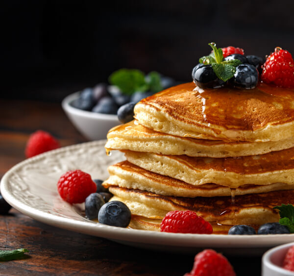 Stos naleśników polanych syropem klonowym, ozdobionych świeżymi jagodami, malinami i liśćmi mięty, serwowany na białym talerzu.