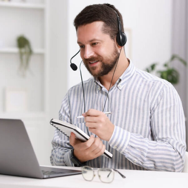 Mężczyzna w biało-szarej koszuli w paski siedzący przy biurku z laptopem, noszący słuchawki z mikrofonem i trzymający notatnik.