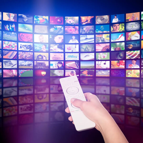 Osoba trzymająca pilot przed ekranem wyświetlającym wiele małych obrazków, reprezentujących różnorodne kanały telewizyjne lub treści multimedialne.