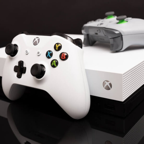 Biała konsola Xbox One S z dwoma białymi kontrolerami na ciemnym tle.