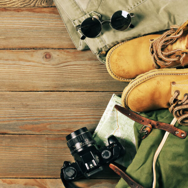 Zestaw przedmiotów turystycznych na drewnianej powierzchni, w tym żółte buty trekkingowe, okulary przeciwsłoneczne, mapa, stary aparat fotograficzny i nóż składany.