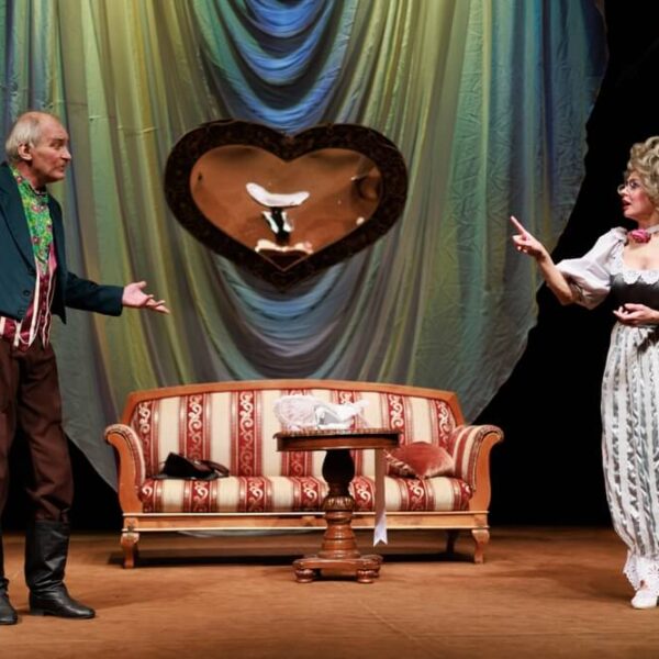 Scena teatralna z dwoma aktorami w kostiumach z epoki; mężczyzna w surducie i kobieta w sukni, stoją po przeciwnych stronach stylowej kanapy na tle dekoracji z dużym, ozdobnym lustrem w kształcie serca.