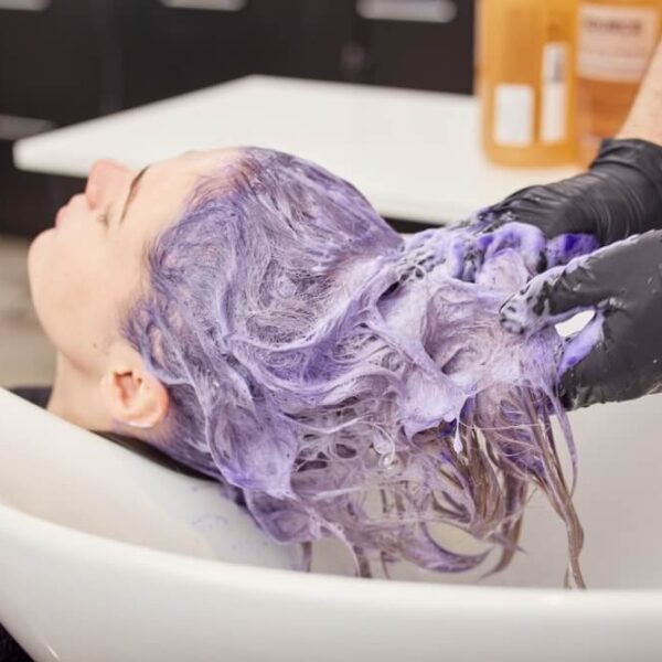 Osoba z fioletową farbą na włosach w salonie fryzjerskim, leżąca przy umywalce, podczas gdy fryzjer w rękawiczkach myje jej włosy.