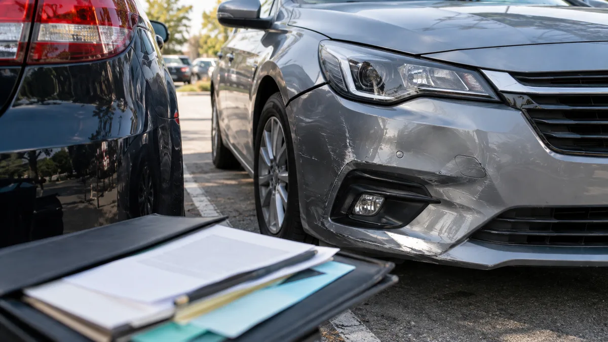 Samochód osobowy po drobnej stłuczce na parkingu z lekko wgniecionym zderzakiem, obok drugi samochód; na pierwszym planie teczka z dokumentami…